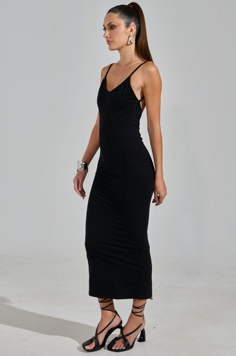 A woman models the MY GOODIES SEAMLESS MAXI DRESS in black, showcasing its bodycon silhouette. She stands in profile with her long hair in a ponytail, accessorized with black strappy sandals and a silver bracelet against a neutral background.