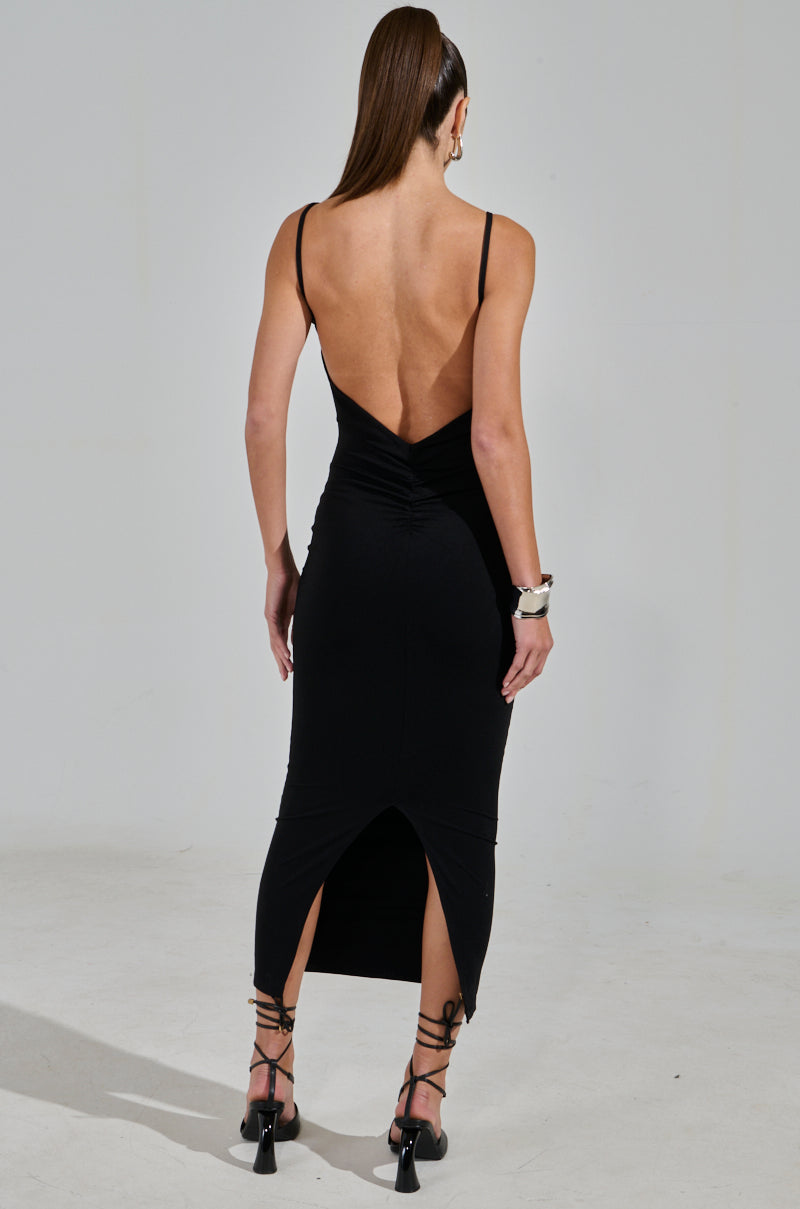 A woman with long hair wears the MY GOODIES SEAMLESS MAXI DRESS, a sleek black bodycon silhouette extending to her calves. The dress features thin straps and a back slit, paired with strappy heels and gold hoop earrings, exuding elegance against a plain gray background.