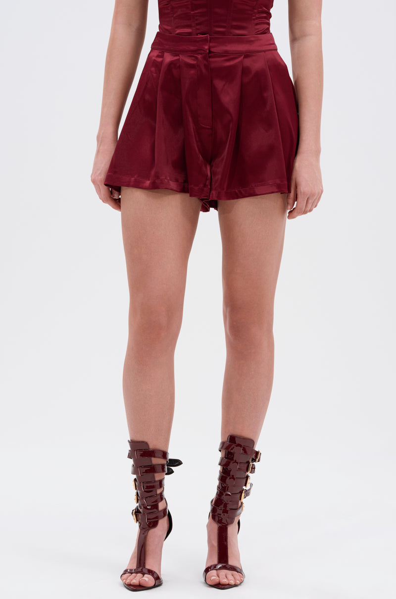 A person wearing the RED HOT FLOUNCY SHORT, a burgundy satin romper with lux shorts, high-rise fit, and flared silhouette, paired with gladiator-style heeled sandals featuring multiple straps and buckles, stands against a white background. 1