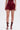 A person wearing the RED HOT FLOUNCY SHORT, a burgundy satin romper with lux shorts, high-rise fit, and flared silhouette, paired with gladiator-style heeled sandals featuring multiple straps and buckles, stands against a white background.