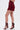A person stands against a white background wearing the RED HOT FLOUNCY SHORT in rich burgundy satin with matching strappy high-heeled sandals featuring multiple gold buckles.