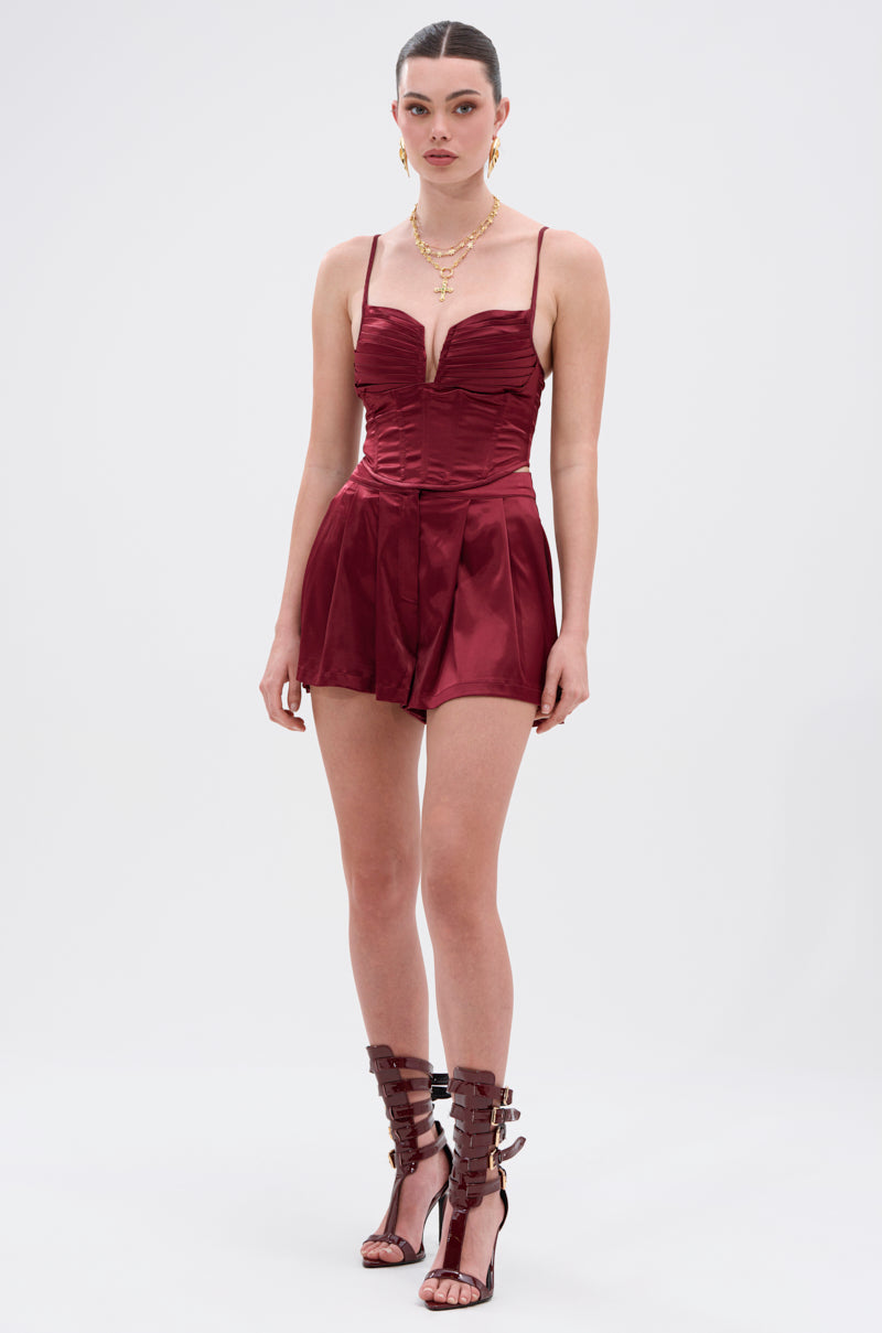 A woman poses against a plain background in the RED HOT FLOUNCY SHORT, a deep burgundy romper with pleated, high-rise fit shorts, styled with layered gold necklaces and strappy brown high-heeled sandals. 4