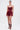 A woman poses against a plain background in the RED HOT FLOUNCY SHORT, a deep burgundy romper with pleated, high-rise fit shorts, styled with layered gold necklaces and strappy brown high-heeled sandals.