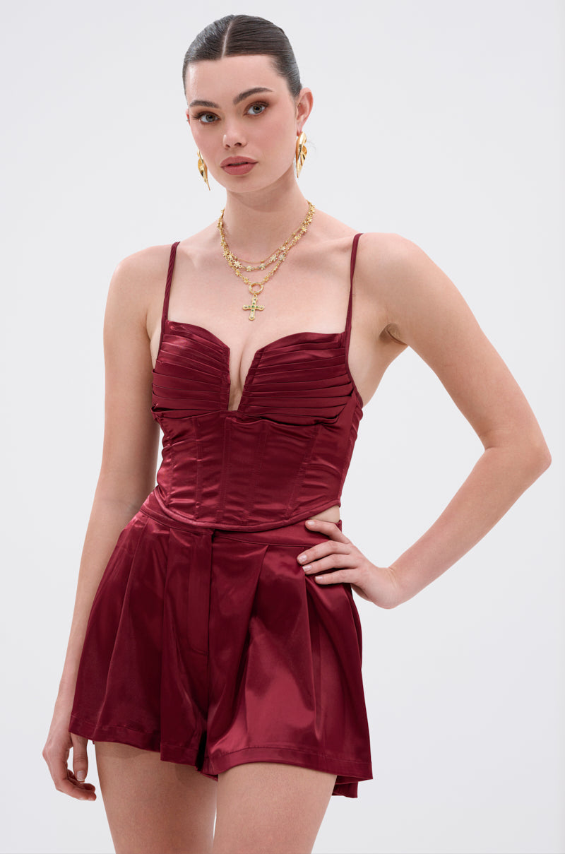 A woman poses confidently in the RED HOT CORSET—a sleeveless burgundy satin romper with a structured bodice and pleated shorts—styled with layered gold necklaces, earrings, and slicked-back hair. 1