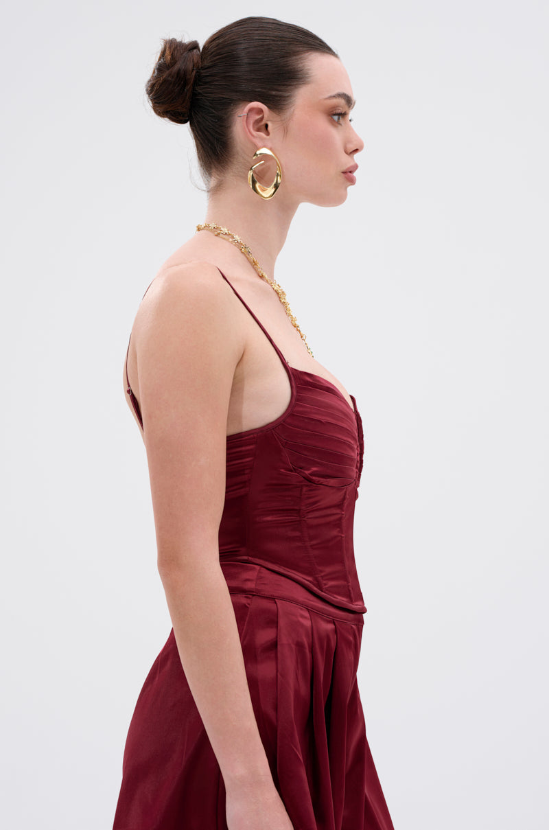 A woman with dark hair in a bun models the RED HOT CORSET, a sleeveless, ruched burgundy satin dress. She accessorizes with large hoop earrings and a gold necklace, posed in profile against a plain light background. 2