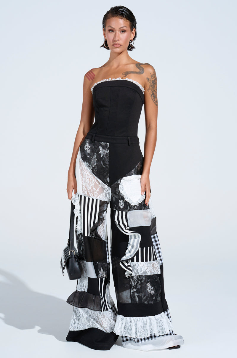 A woman with short hair wears the IRIS PATCHWORK JUMPSUIT, a black strapless jumpsuit with wide-leg patchwork details. She carries a small black handbag and has visible tattoos on her arms and shoulders.