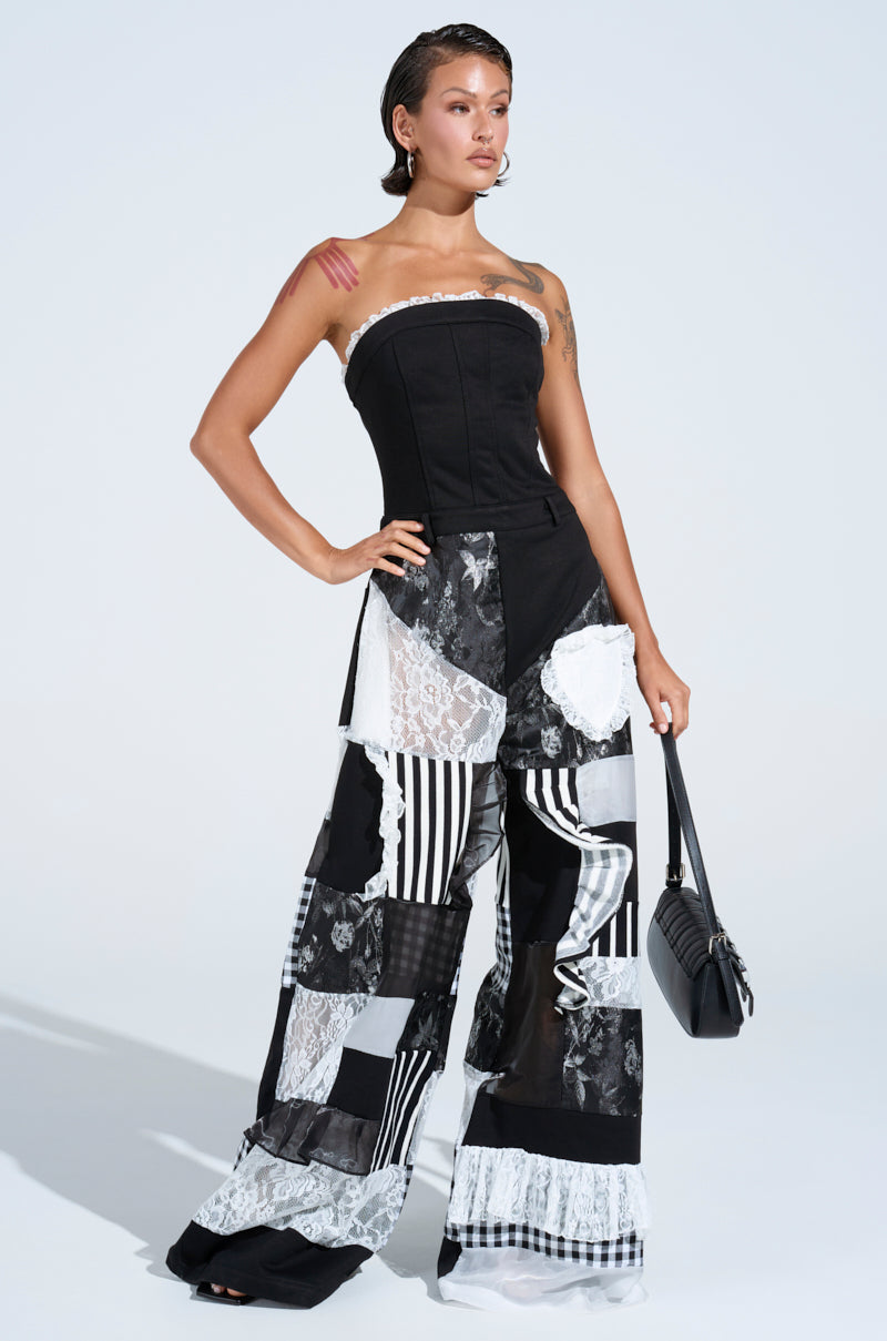 A woman with short, slicked-back hair wears the IRIS PATCHWORK JUMPSUIT, a strapless design featuring black, white, lace, and striped patterns. She pairs it with a black handbag and poses confidently against a plain light background.