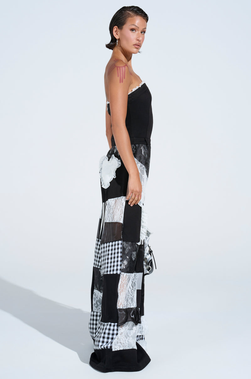 A woman in profile wears the IRIS PATCHWORK JUMPSUIT, a black strapless design with a lace and checkered patchwork skirt. She has short, slicked-back hair, pink marks on her right shoulder, and stands against a plain white background.