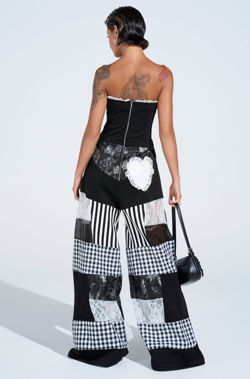 A person with short slicked-back hair, tattoos, and a strapless black top faces away, wearing the IRIS PATCHWORK JUMPSUIT: black-and-white wide-leg pants with lace and checkered patterns, and holding a small black handbag.