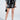 Wearing MY LIFE CROC SHORT, a person in a wavy black-and-white striped top holds a small black purse and wears black and gold high heels. A small tattoo above their ankle adds character against the minimalist background.