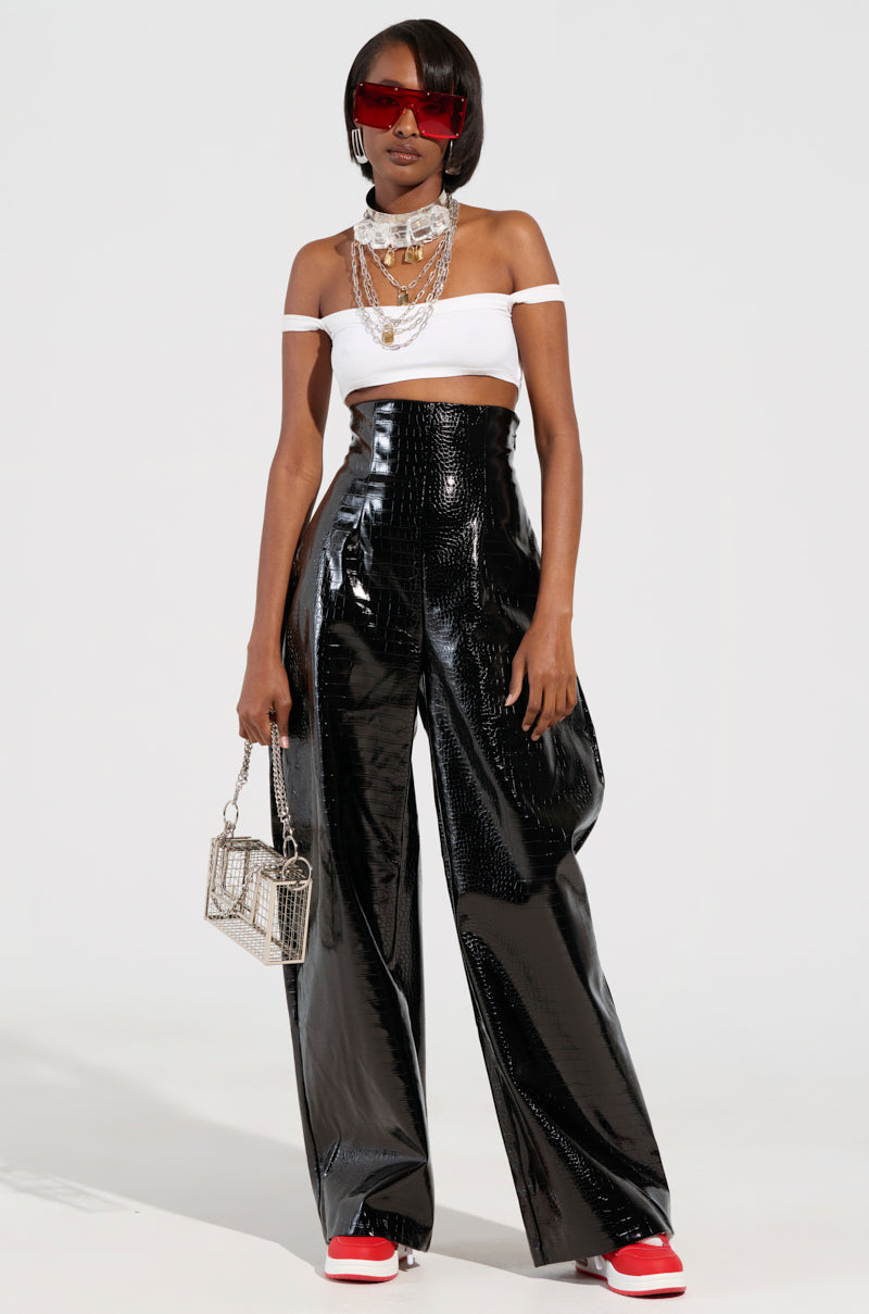 A fashionable individual dons an off-shoulder white top paired with MY LIFE CROC TROUSER—faux patent leather vinyl pants featuring a high-rise waistband. They complete the look with red sneakers and oversized red sunglasses, while holding a small rectangular purse and wearing layered necklaces, striking a confident pose against a plain background.