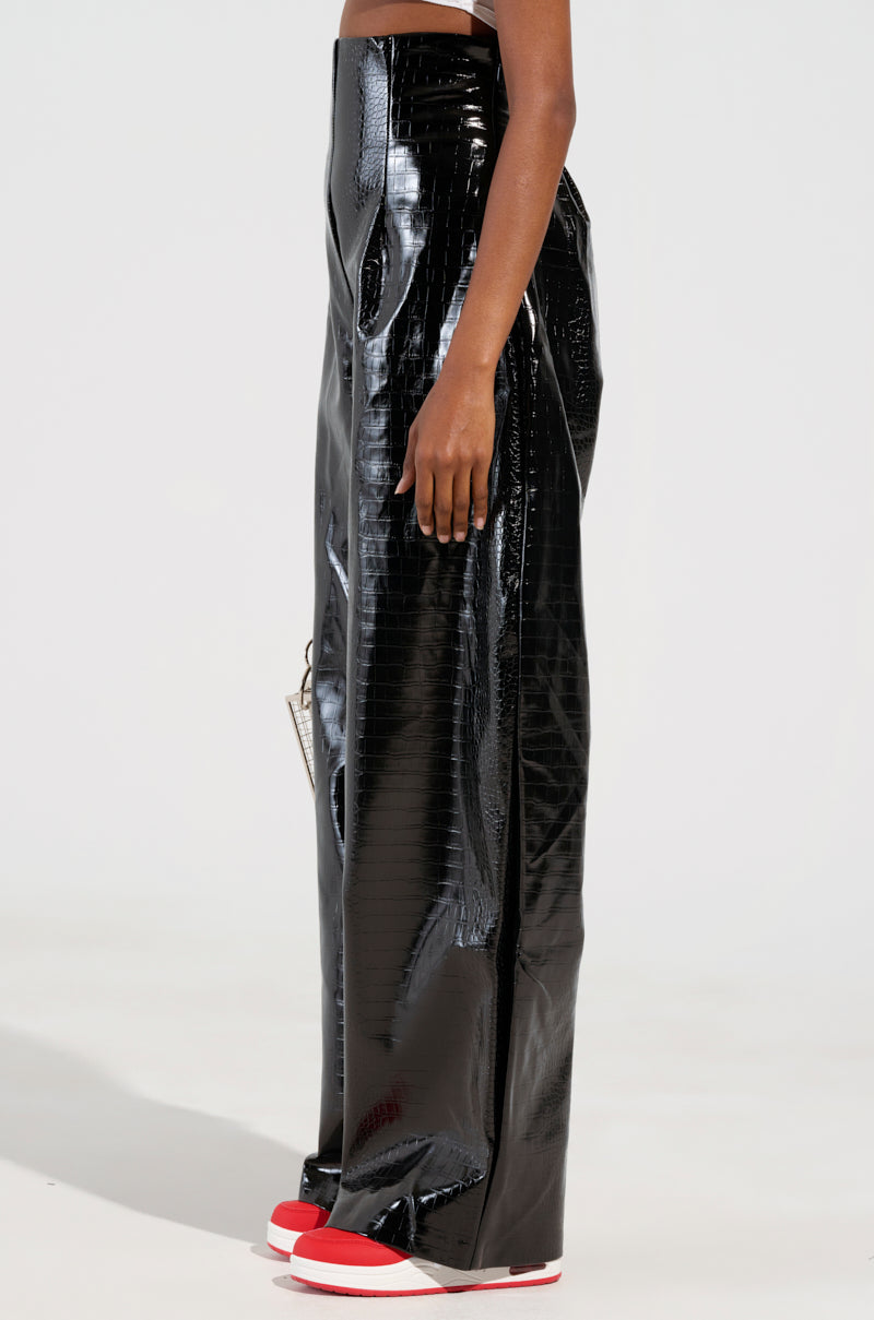 A person wearing MY LIFE CROC TROUSER in high-waisted, faux patent leather black vinyl stands in profile against a plain background. The crocodile-textured pants are paired with white sneakers featuring red soles, creating a sleek and stylish look.