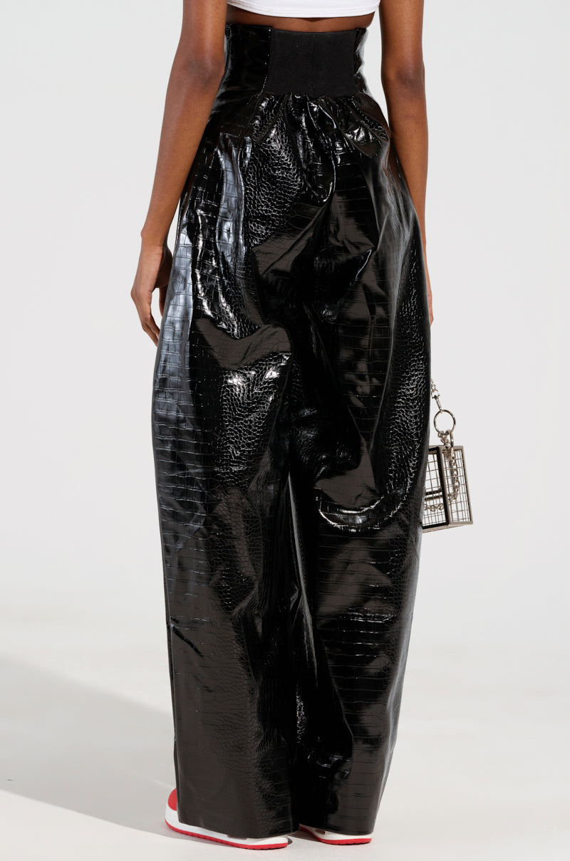 A person is wearing the MY LIFE CROC TROUSER, which features a shiny black wide-leg design with a faux patent leather texture, alongside a white top and red-soled shoes. They are carrying a small cage-like handbag against a plain, light-colored background.