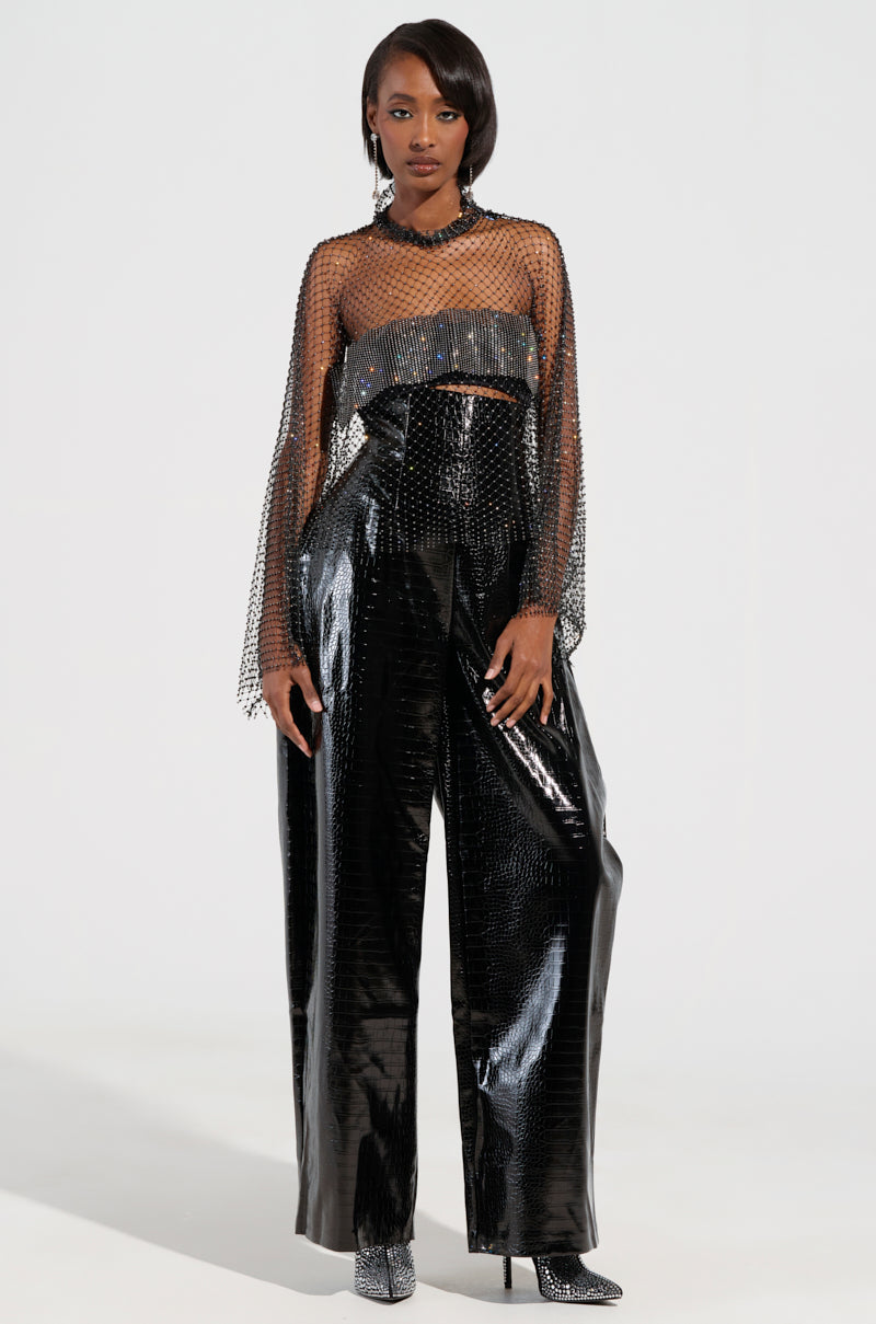 A person is wearing a trendy, sheer top with long sleeves paired with the MY LIFE CROC TROUSER, shiny black pants that have a high waist and a wide-leg silhouette. Silver accessories and ankle-length boots complete this sleek and futuristic look.
