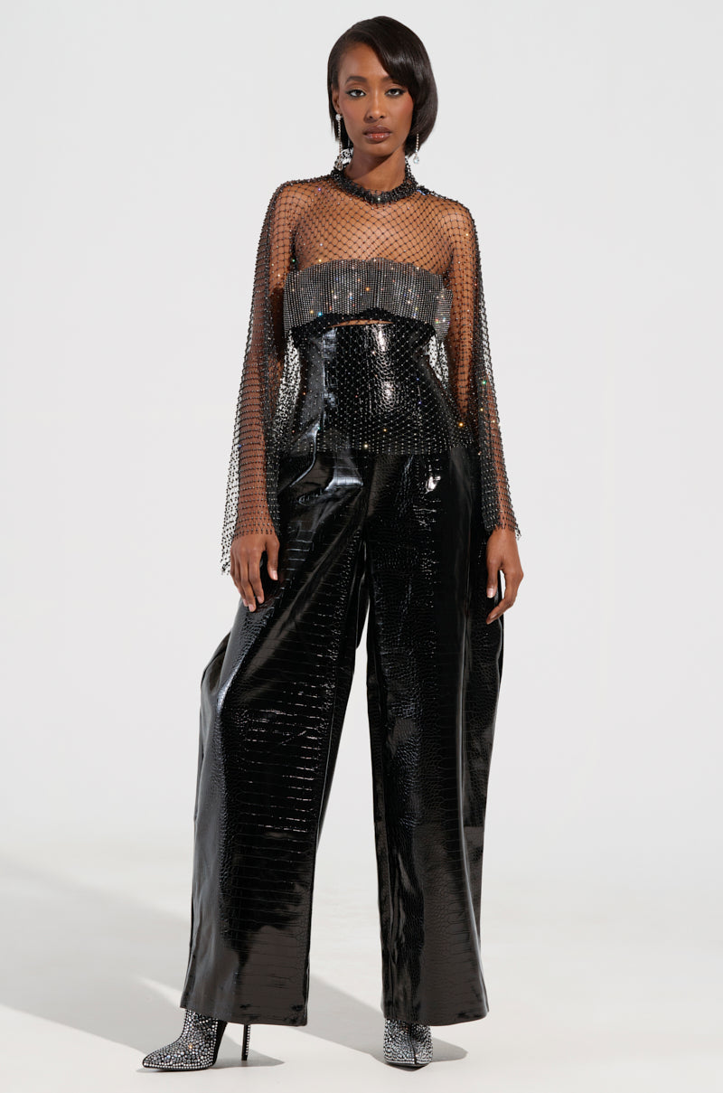 A person wearing a stylish outfit, featuring a sheer, beaded top and the MY LIFE CROC TROUSER in black faux patent leather with wide legs, stands confidently. The high-rise waistband complements their high heels, matching the shimmering details of the ensemble against the plain white background.