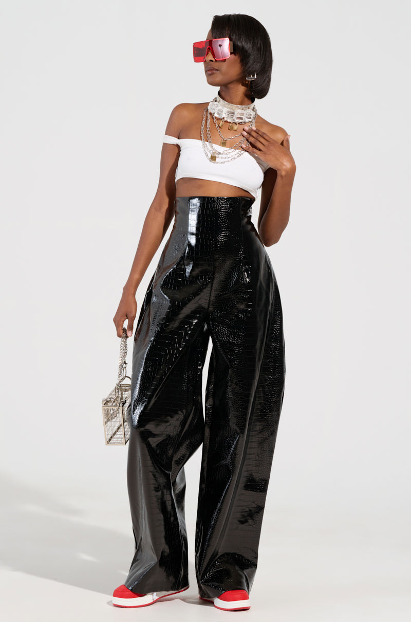 A person poses stylishly in MY LIFE CROC TROUSER, featuring an oversized black faux patent leather design with a wide-leg silhouette, paired with a white cropped top, chunky white sneakers, red sunglasses, and layered silver necklaces while holding a small patterned handbag.