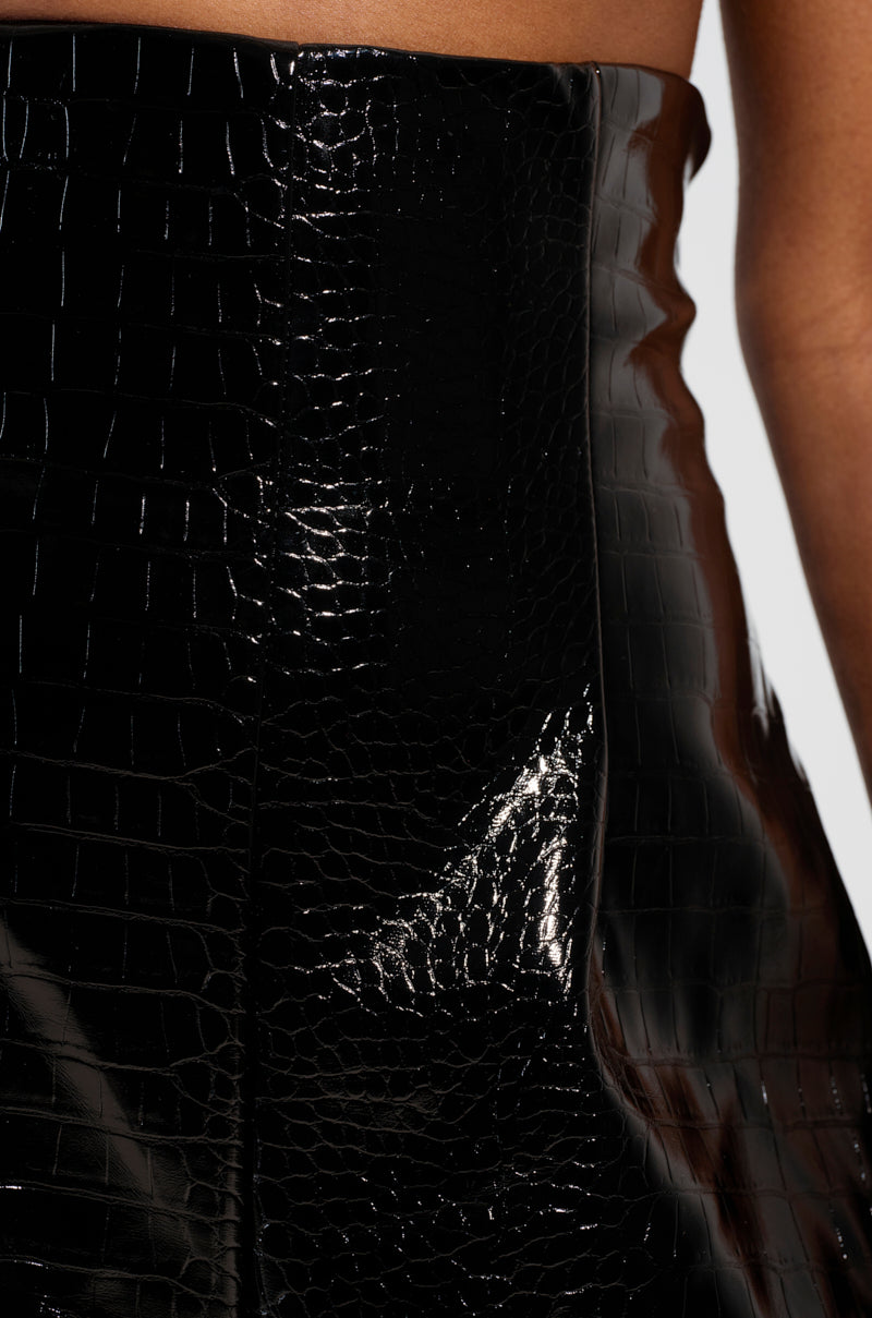 A close-up of the shiny black faux patent leather fabric of the MY LIFE CROC TROUSER showcases its smooth texture. The material reflects light, accentuating its glossy finish and intricate crocodile pattern design.