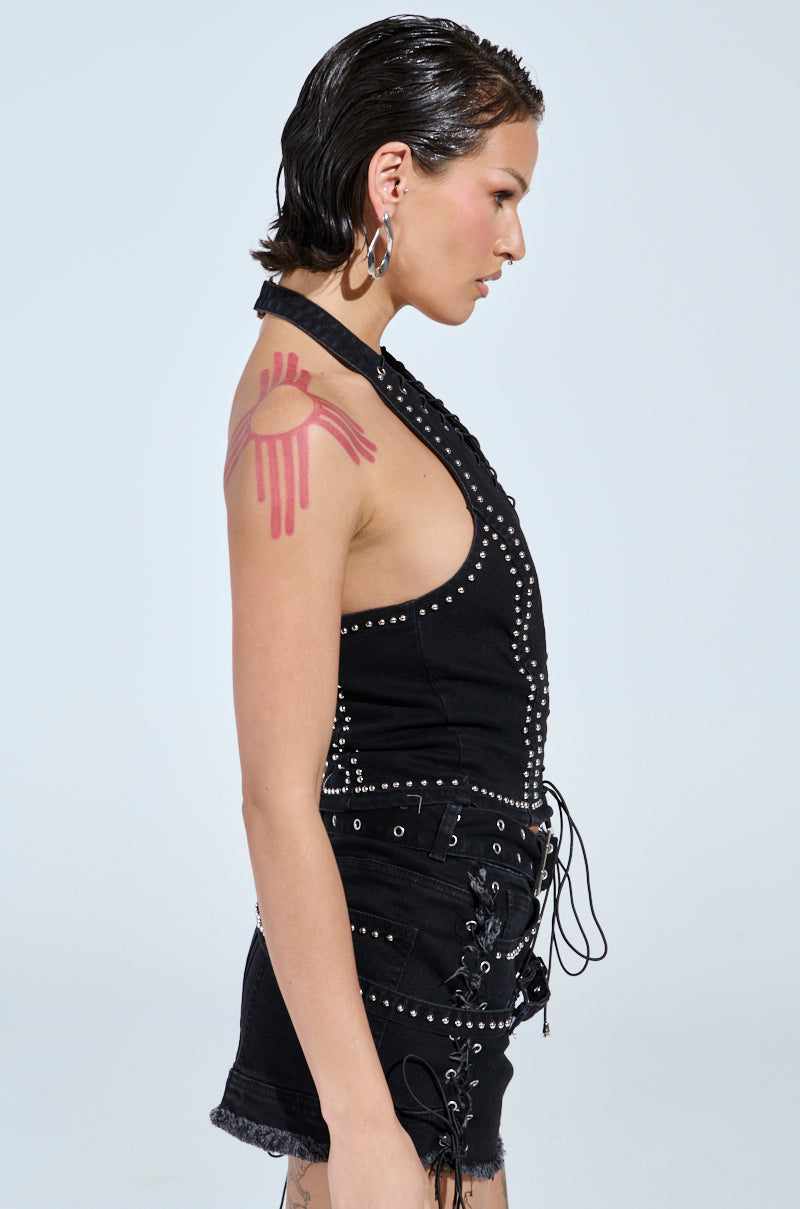 A person with short dark hair wears the CATCH ME AFTER MIDNIGHT STUDDED CORSET TOP and matching shorts, featuring metallic studs and lace-up details. They display a pink shoulder tattoo and large hoop earrings, posing in profile against a plain background.