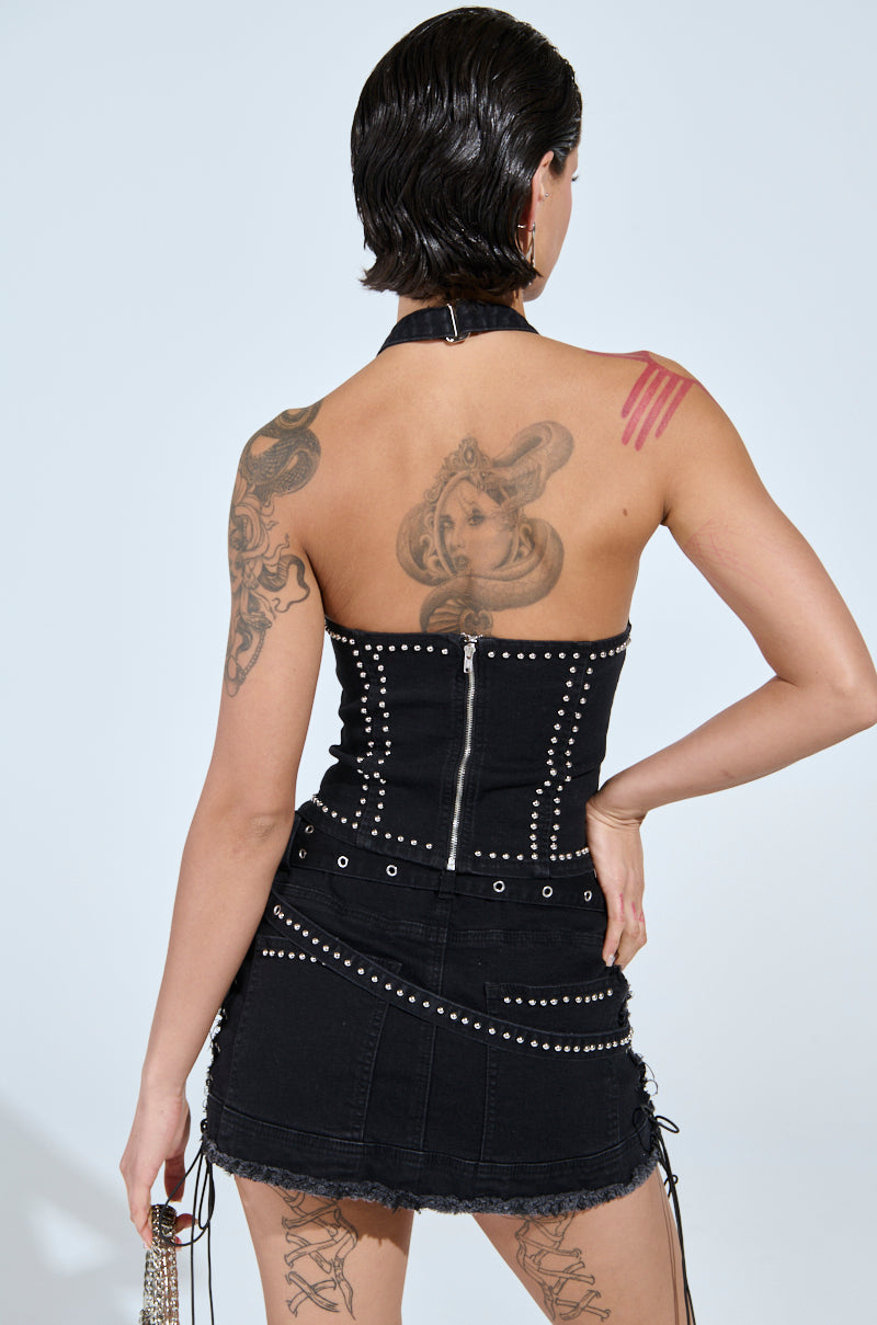A person with short hair is seen from the back, wearing the CATCH ME AFTER MIDNIGHT STUDDED CORSET TOP and a skirt adorned with lace trim and grommets, tied at the sides. Tattoos grace their back and shoulder against a light gray backdrop.