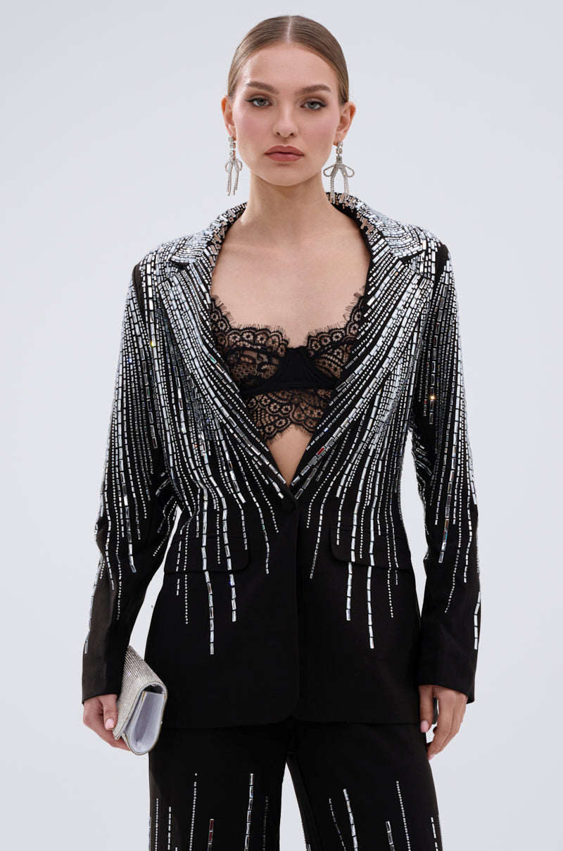 A woman with slicked-back hair models the MILA EMBELLISHED BLAZER IN BLACK with silver sequin stripes, a black lace bralette, matching stretch pants, dangling earrings, and a silver clutch against a plain backdrop.