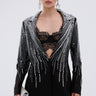 A woman with slicked-back hair models the MILA EMBELLISHED BLAZER IN BLACK with silver sequin stripes, a black lace bralette, matching stretch pants, dangling earrings, and a silver clutch against a plain backdrop.
