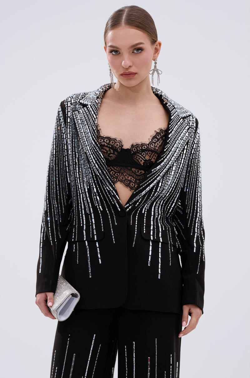 A woman in the MILA EMBELLISHED BLAZER IN BLACK with matching pants and silver sequin stripes over a lacy black bralette, holding a small silver clutch. She wears her hair slicked back, with neutral makeup and statement earrings.