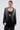 A woman in the MILA EMBELLISHED BLAZER IN BLACK with matching pants and silver sequin stripes over a lacy black bralette, holding a small silver clutch. She wears her hair slicked back, with neutral makeup and statement earrings.