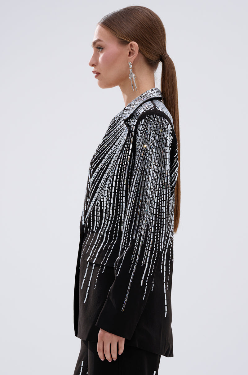 A woman with long brown hair in a low ponytail wears the MILA EMBELLISHED BLAZER IN BLACK with silver bead fringe and matching dangling earrings, shown in profile against a plain light background.