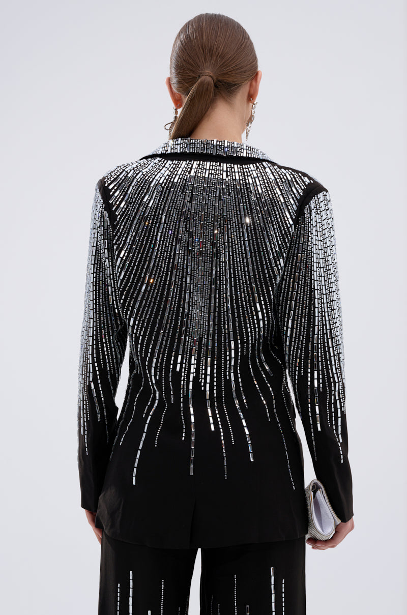 A person with straight brown hair in a low ponytail is seen from behind, wearing the MILA EMBELLISHED BLAZER IN BLACK adorned with vertical rows of silver sequins and holding a silver clutch.