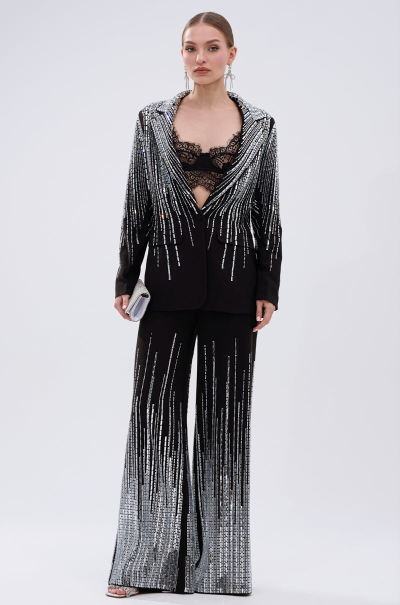 A woman stands confidently against a light background wearing the MILA EMBELLISHED BLAZER IN BLACK with silver sequin stripes, paired with a lace bralette, statement earrings, metallic sandals, and a small white clutch.
