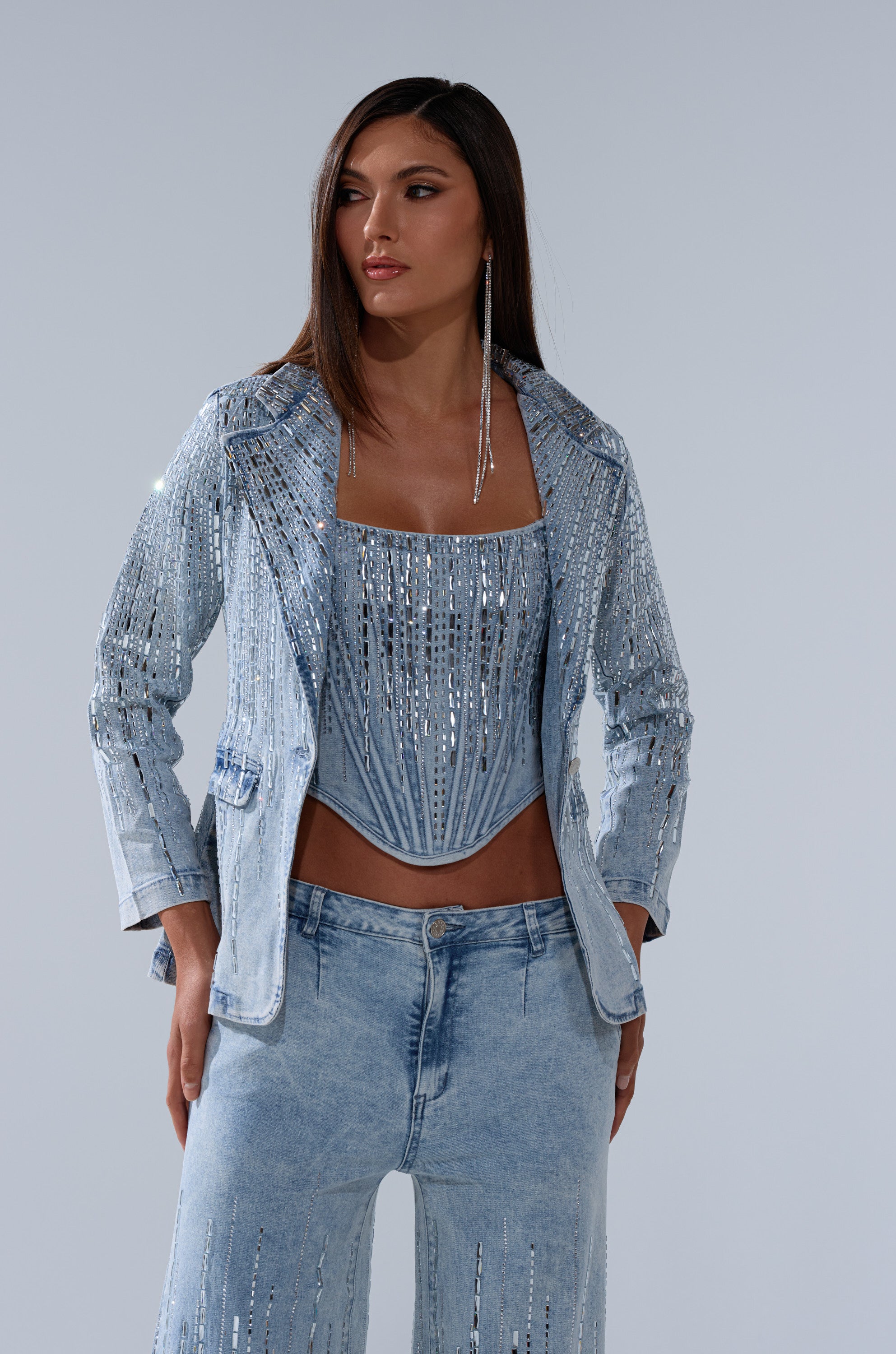 A woman wearing the MILA EMBELLISHED BLAZER IN DENIM, styled with high-waisted jeans and a rhinestone corset top, stands against a plain background, looking to her left.
