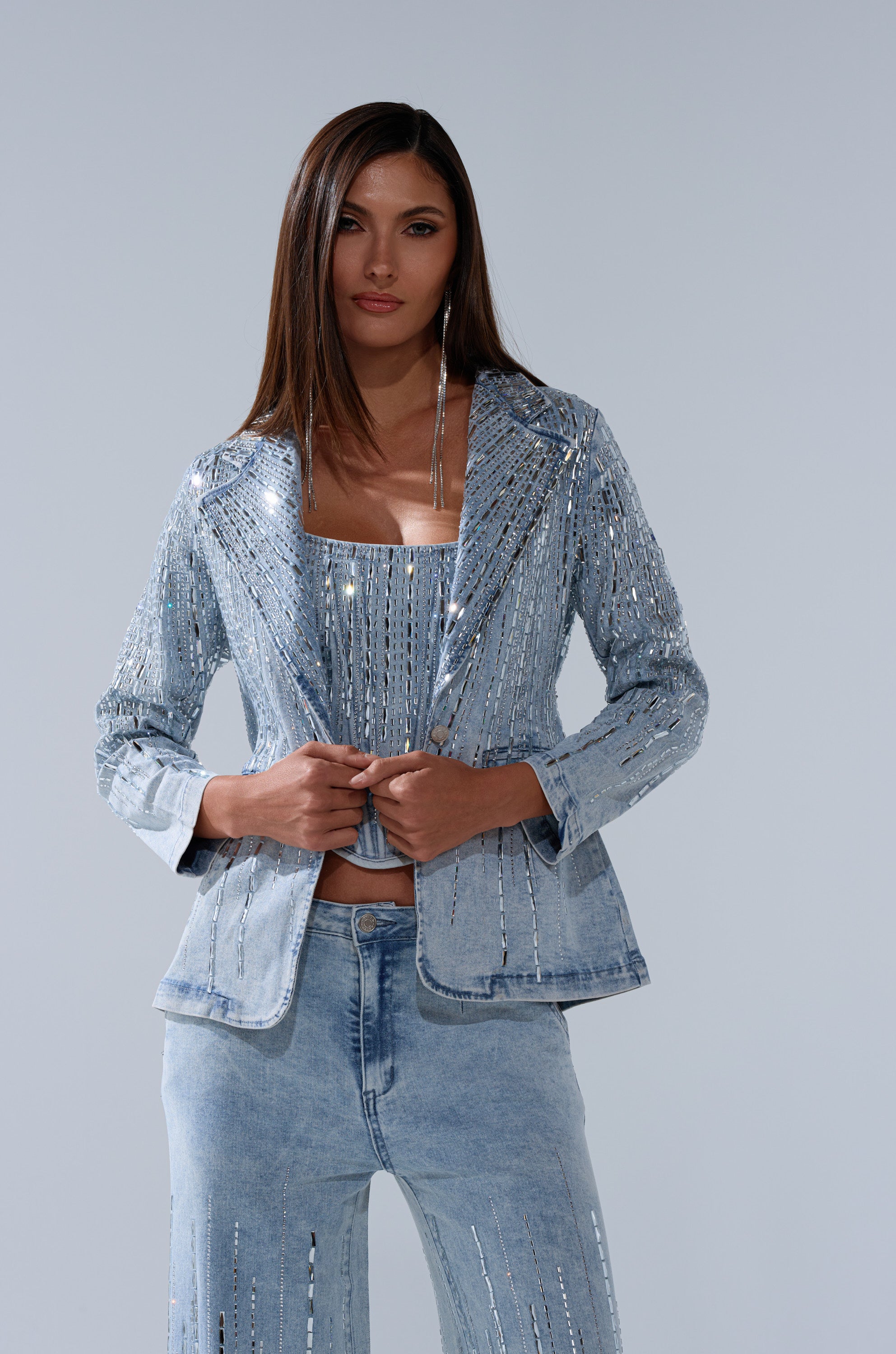 A woman with long, straight brown hair confidently poses in the MILA EMBELLISHED BLAZER IN DENIM, featuring sparkling silver embellishments, paired with a light blue suit against a light gray background.