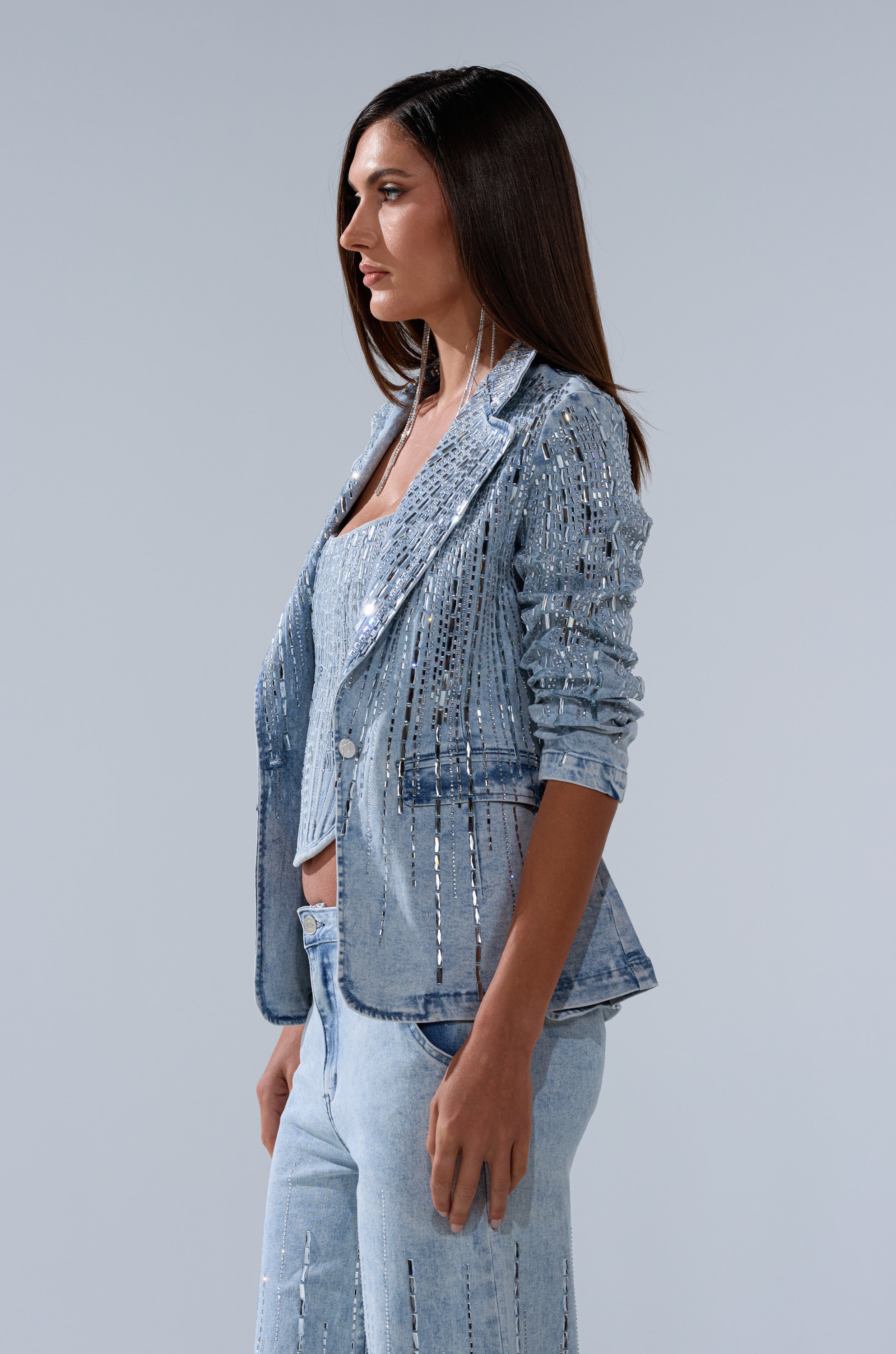 A woman with straight dark hair stands in profile against a light gray background, wearing the MILA EMBELLISHED BLAZER IN DENIM from AKIRA Label, styled with a matching top and jeans featuring rhinestone accents.