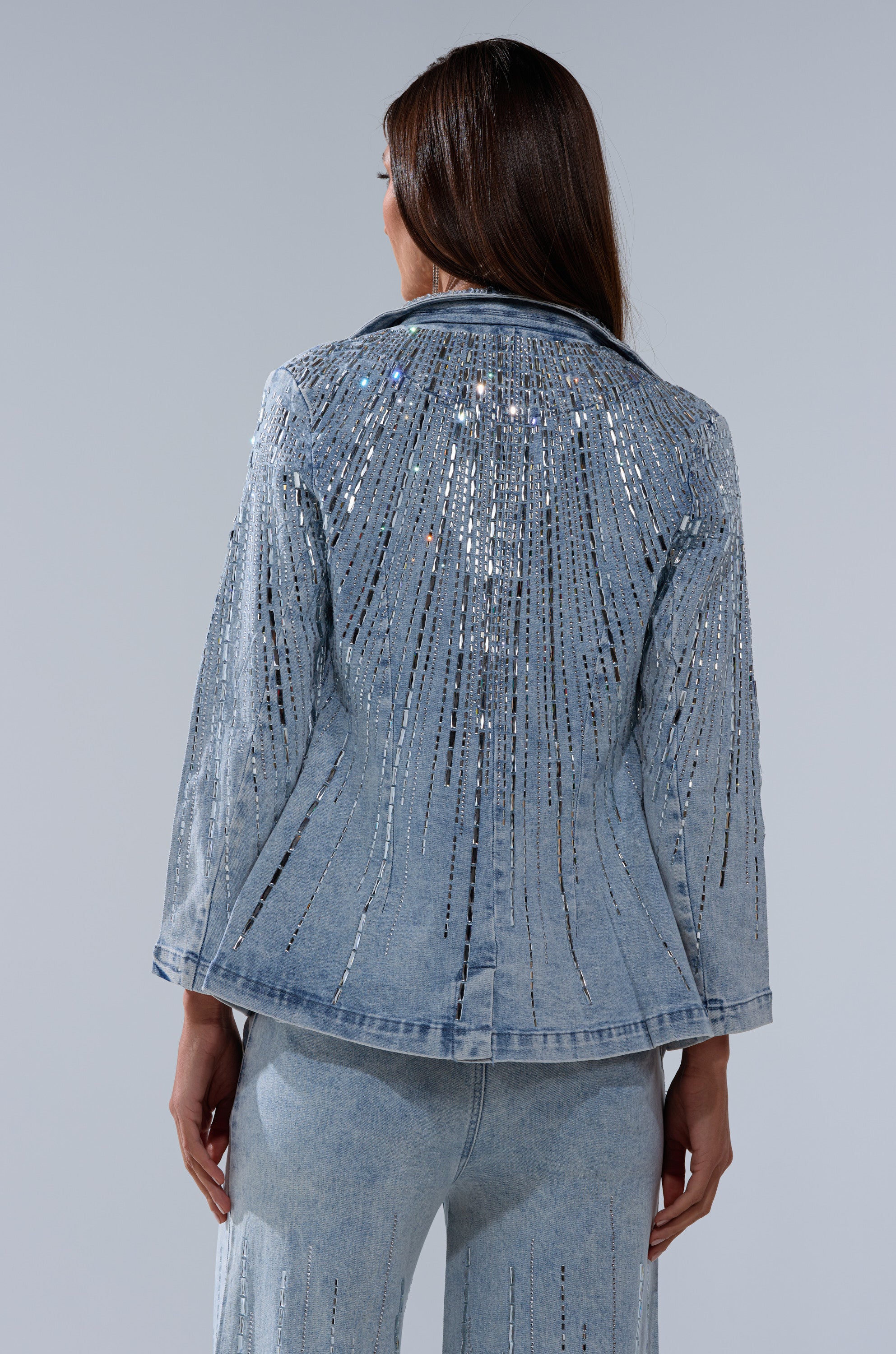 A woman faces away from the camera, wearing the MILA EMBELLISHED BLAZER IN DENIM. This AKIRA Label jacket features vertical rhinestone detailing for a radiant look, paired with matching pants.