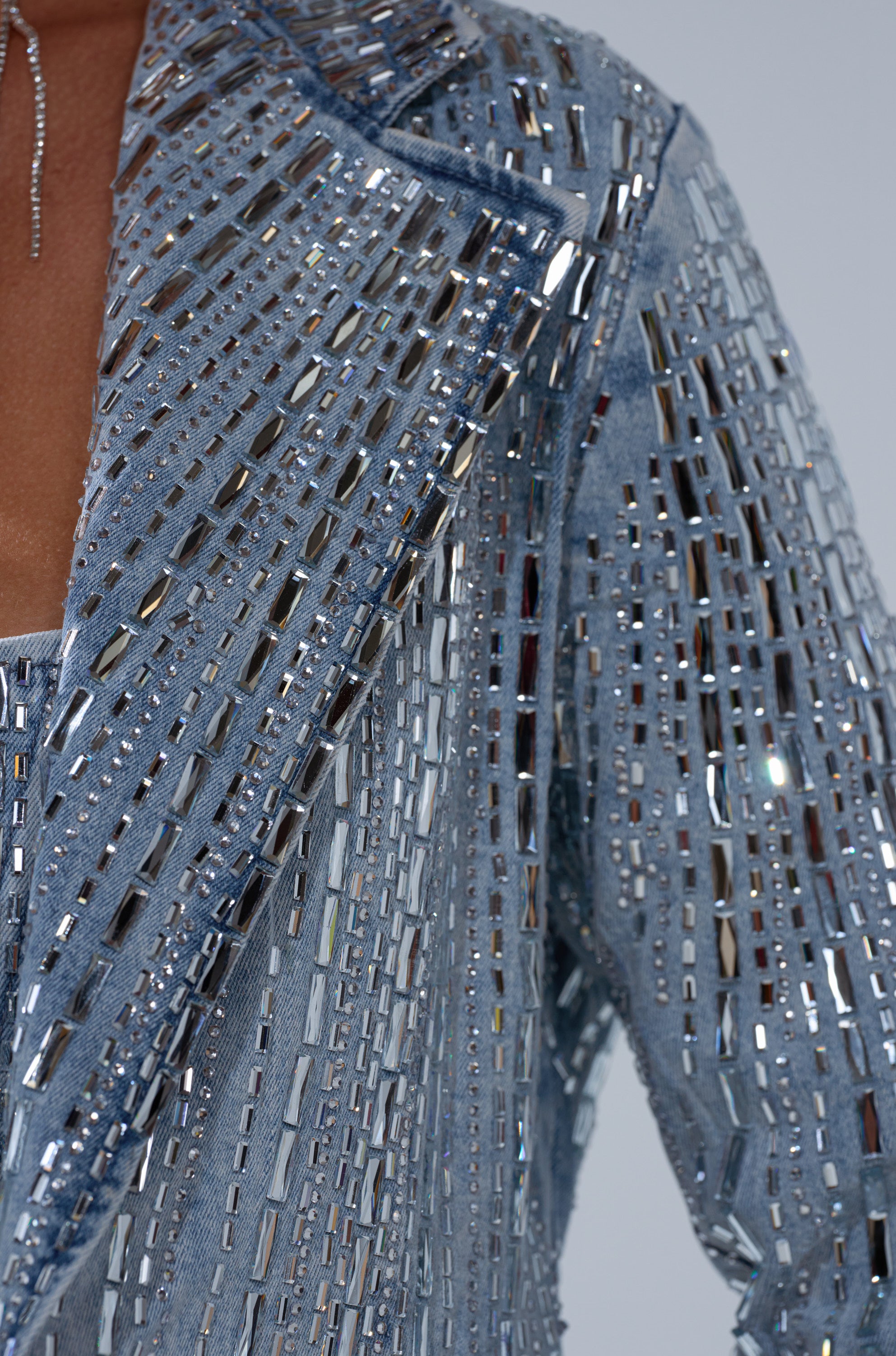 Close-up of the MILA EMBELLISHED BLAZER IN DENIM by AKIRA Label, featuring blue denim covered in rows of rectangular silver and clear rhinestones for a sparkling effect, worn over a matching top.
