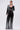 A woman poses in the MILA EMBELLISHED FLARE PANT IN BLACK, featuring vertical silver sequin stripes. She wears a black lace bralette, holds a silver clutch, and stands against a plain light background.