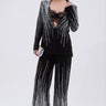 A woman poses in the MILA EMBELLISHED FLARE PANT IN BLACK, featuring vertical silver sequin stripes. She wears a black lace bralette, holds a silver clutch, and stands against a plain light background.