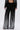 A person wears the MILA EMBELLISHED FLARE PANT IN BLACK—high-rise, wide-leg pants with vertical silver rhinestones—paired with a matching jacket and holding a small silver clutch. The photo is cropped at the waist and feet.