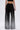 A person is shown from the back wearing the MILA EMBELLISHED FLARE PANT IN BLACK featuring sparkling rhinestones near the hem, paired with a black crop top and holding a silver clutch.