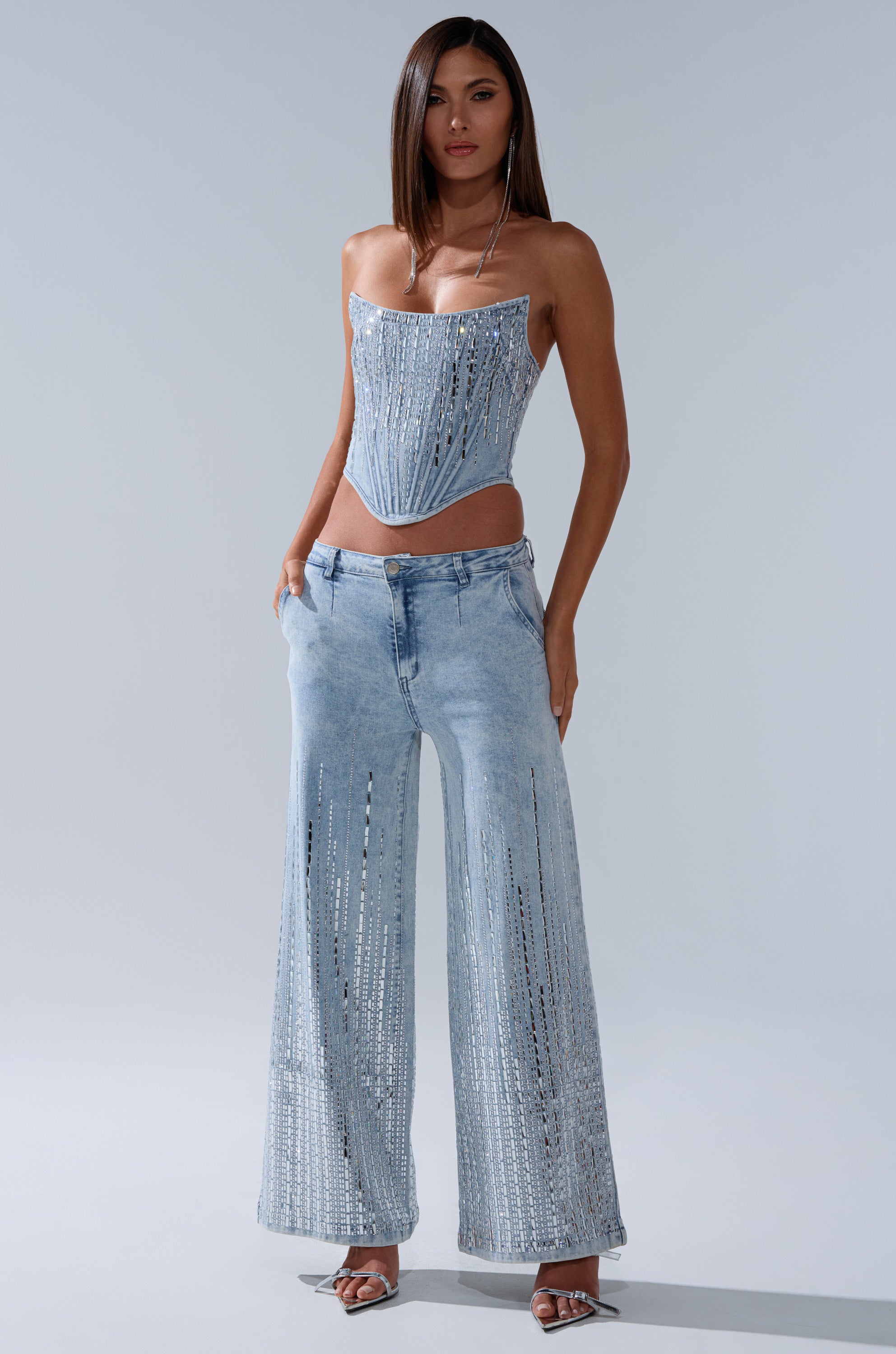 A woman faces forward in the MILA EMBELLISHED FLARE PANT IN DENIM, featuring high-rise wide legs with vertical rhinestone details. She pairs it with a strapless beaded denim top, sandals, a necklace, and wears her hair straight.