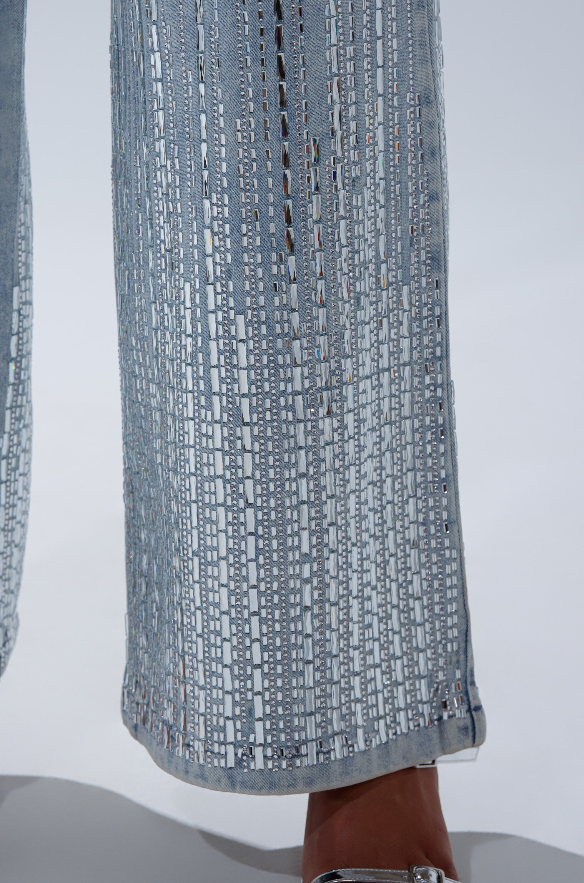 Close-up of a person's lower legs in the MILA EMBELLISHED FLARE PANT IN DENIM, featuring vertical and horizontal cutouts that reveal skin. The pants have a flared silhouette and are styled with silver open-toe shoes.