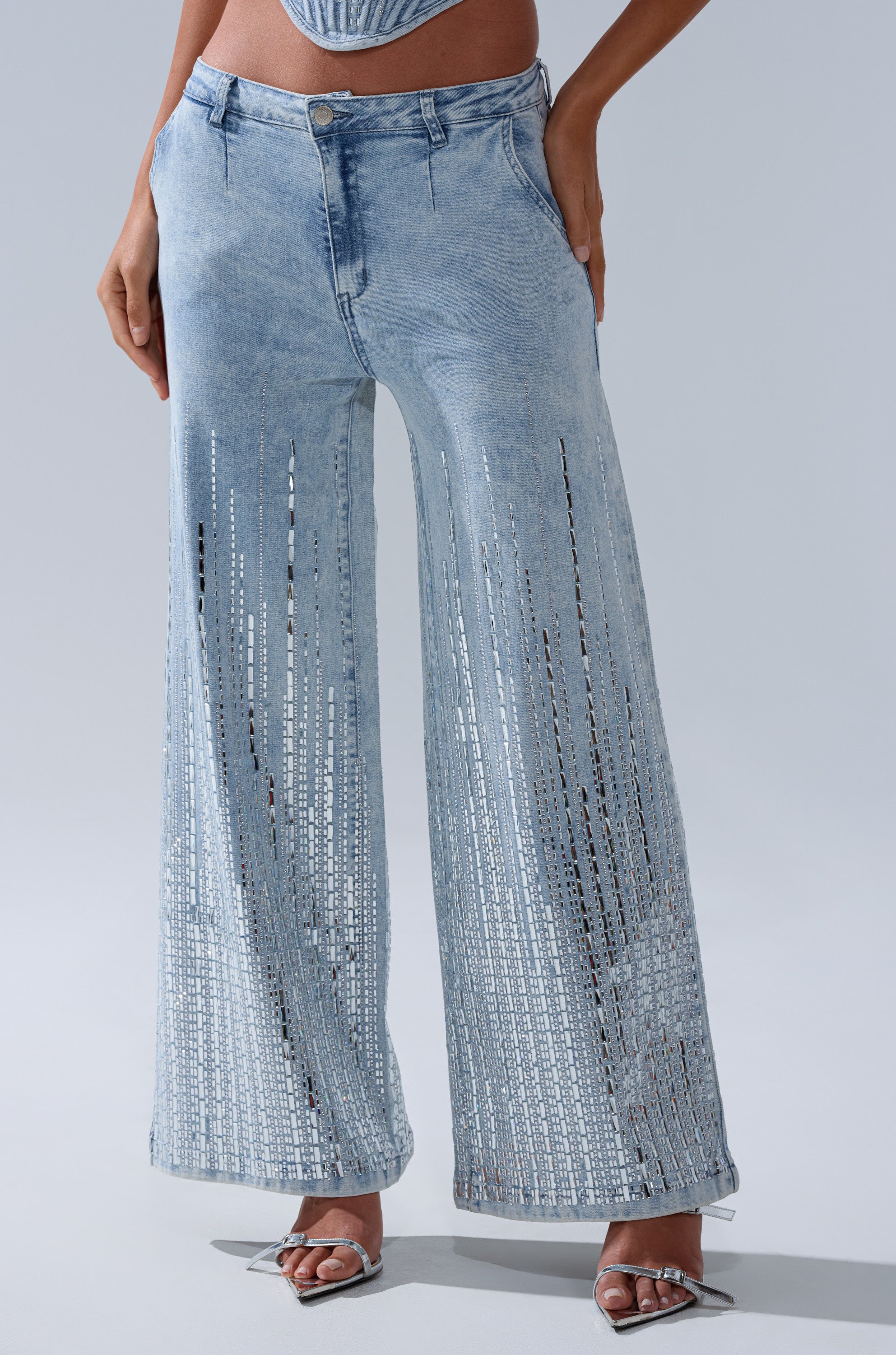 A person wears the MILA EMBELLISHED FLARE PANT IN DENIM with silver high-heeled sandals, standing against a plain light gray background.