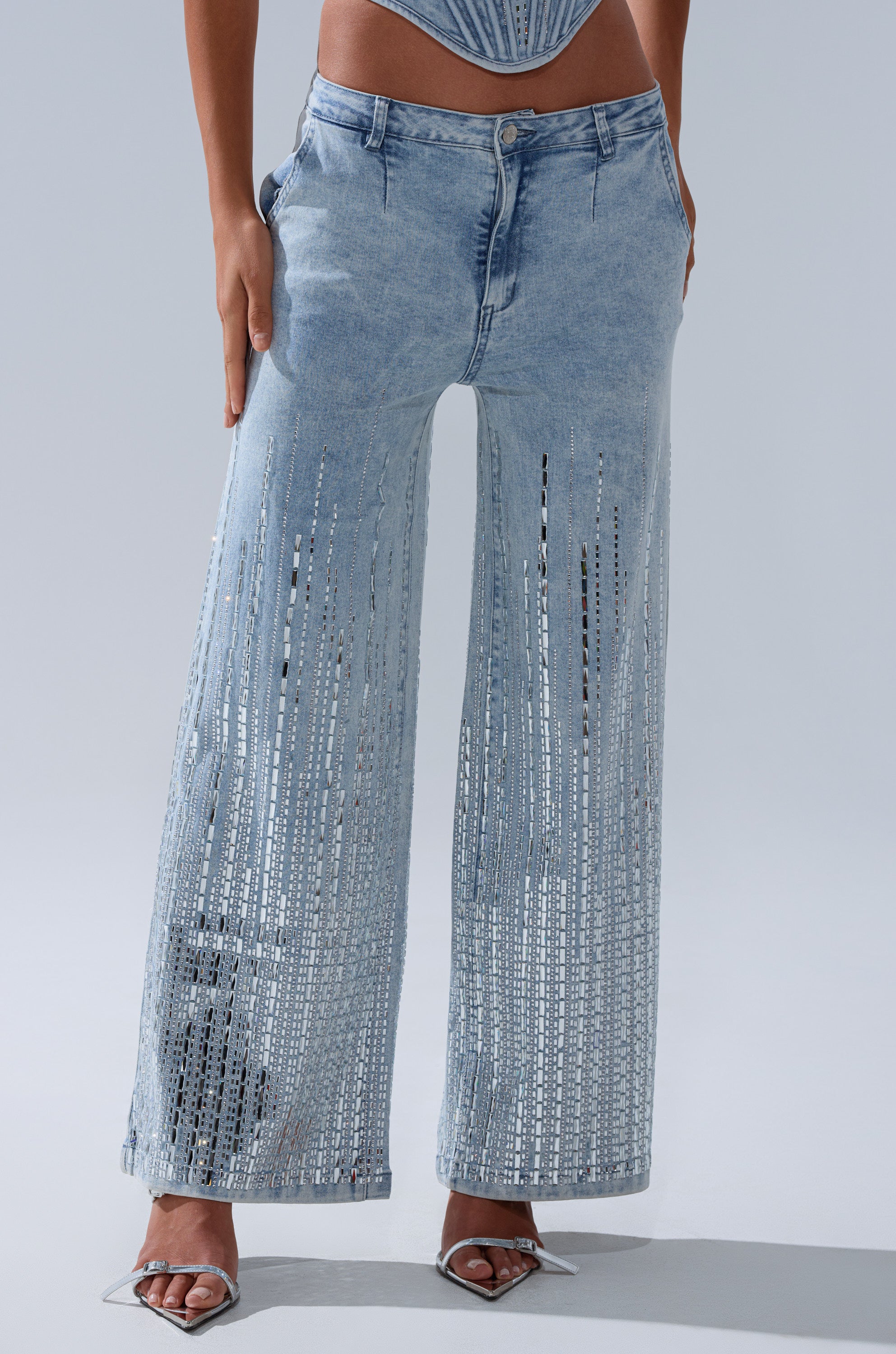A person wears the MILA EMBELLISHED FLARE PANT IN DENIM, featuring high-rise wide legs with vertical slits from thigh to hem that reveal skin. Styled with silver pointed-toe heels. Upper body mostly out of frame.
