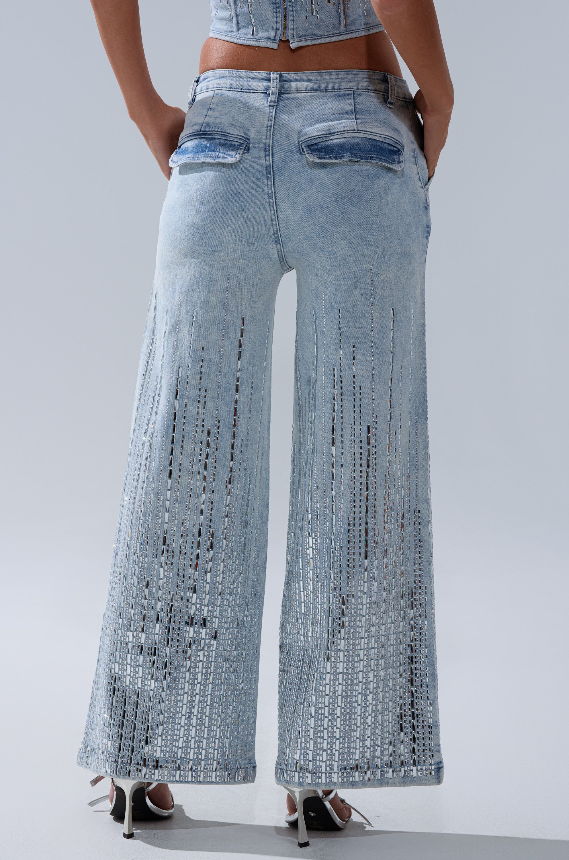 A person wearing the MILA Embellished Flare Pant in Denim, featuring light blue high-rise wide legs with vertical cut-out details, paired with high heels and shown from the back against a plain light background.