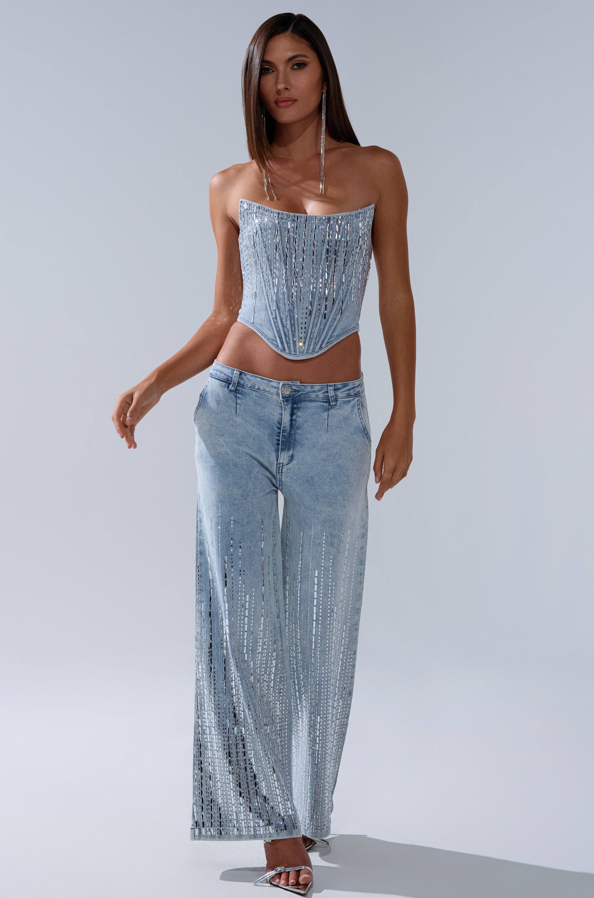 A woman stands against a plain background wearing the MILA EMBELLISHED FLARE PANT IN DENIM, styled with a strapless light blue denim corset top and heeled sandals. She has long straight hair and silver sequin details on her high-rise wide-leg jeans.