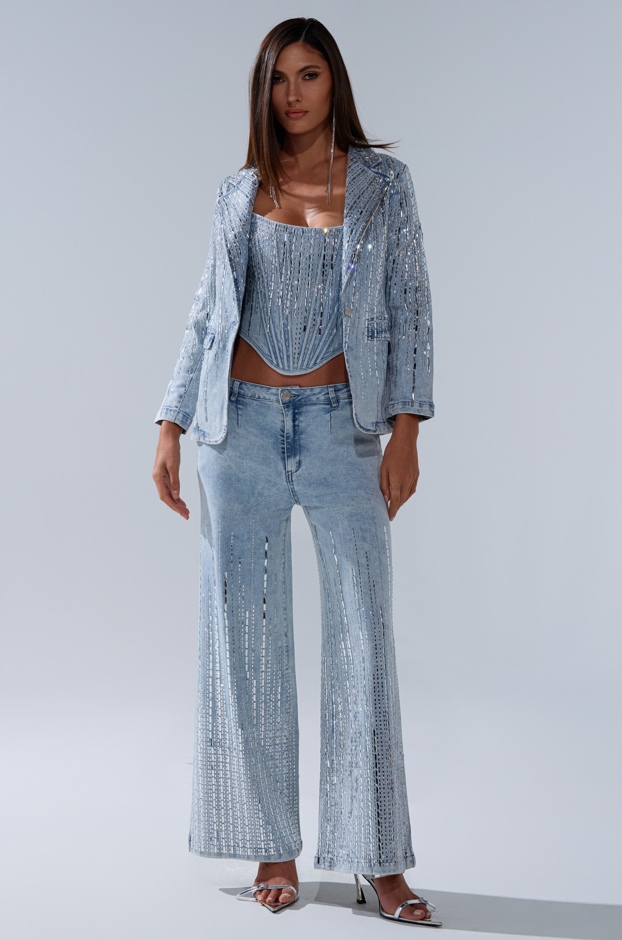 A woman poses confidently in the MILA EMBELLISHED FLARE PANT IN DENIM—a sparkly, light blue rhinestone flare pant—paired with a matching embellished corset top, silver heels, and straight brown hair against a plain background.