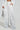 Donning the MILA EMBELLISHED FLARE PANT, a person stands confidently in a white outfit with vertical silver sequin stripes, one hand in their pocket. They hold a matching sparkling clutch, showcasing the high-rise wide-leg pants and jacket with a metallic reflective design.