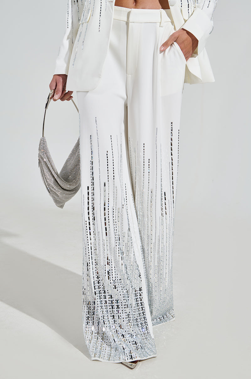 Wearing the MILA EMBELLISHED FLARE PANT with silver sequins, paired with a matching sequined jacket and handbag, along with white heels. This sparkling ensemble stands out against the plain white background.