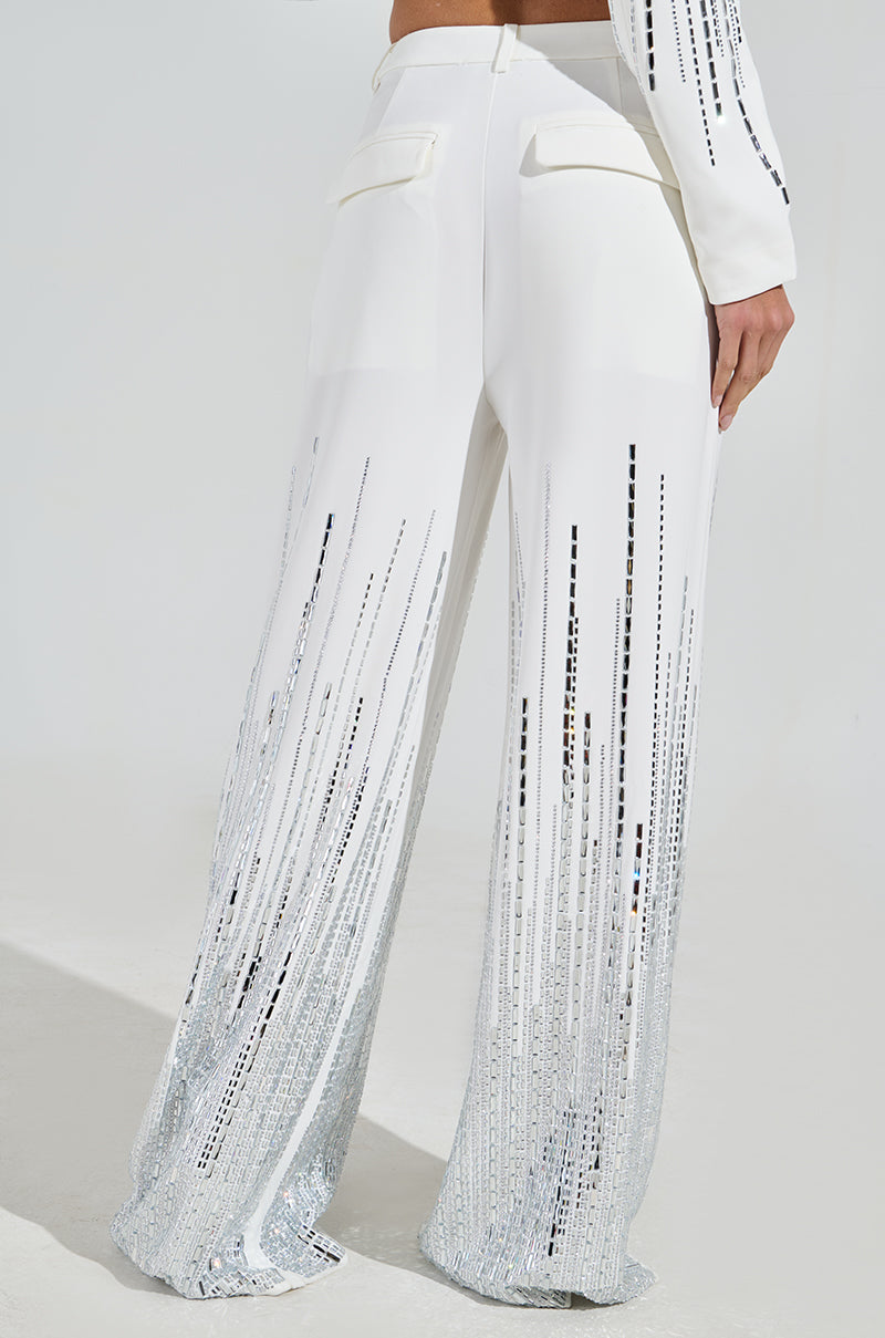 Someone is dressed in the MILA EMBELLISHED FLARE PANT, high-rise wide-leg white pants with sparkling silver stripes from waist to hem, featuring two back pockets. They are paired with a matching long-sleeve top.