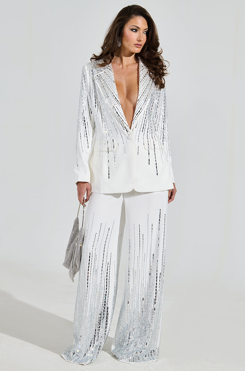 A woman poses against a neutral backdrop in MILA EMBELLISHED FLARE PANTS, adorned with sparkling silver rhinestones. The wide-leg style complements her deep V-neckline top. She holds a matching silver clutch, and her long hair flows over her shoulders.